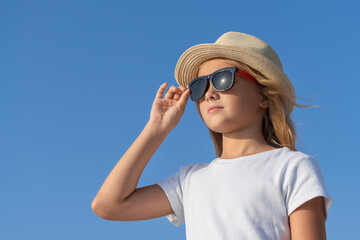 A young girl in a hat and sunglasses adjusts her glasses with one hand.