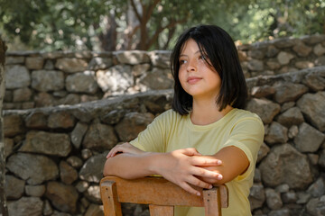 A young teenager girl sits on a wooden chair against a stone wall.