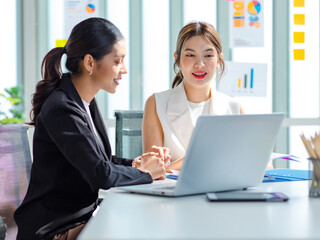 Two millennial Asian young beautiful professional successful businesswoman colleagues sitting smiling in meeting room having funny conversation together while working with laptop notebook computer