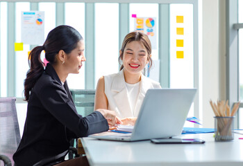 Fototapeta premium Two millennial Asian young beautiful professional successful businesswoman colleagues sitting smiling in meeting room having funny conversation together while working with laptop notebook computer