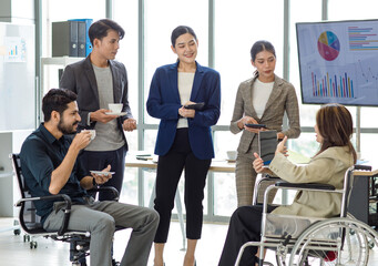 Group of millennial multinational multicultural Asian Indian businessmen and businesswomen with disabled handicapped partner sitting on wheelchair taking coffee break discussing together in office