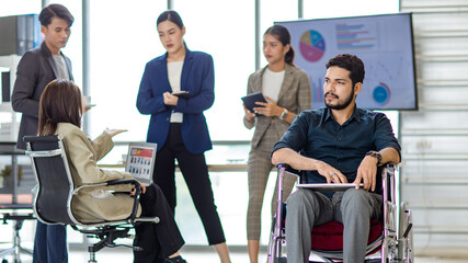 Indian bearded disabled handicapped businessman sitting working with laptop computer on wheelchair while Asian male and female colleagues hold tablet talking discussing together in blurred background
