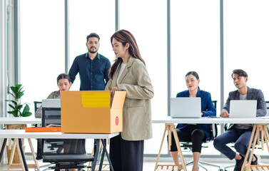 Portrait shot of Asian sad jobless businesswoman in casual suit standing holding belongings in cardboard box after fired while male and female colleagues waving hands goodbye in blurred background