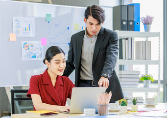 Millennial male businessman mentor standing holding disposable coffee cup teaching coaching Asian intern trainee secretary working with laptop computer in multinational company office