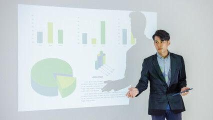 Millennial Asian young cheerful male businessman presenter in formal suit standing holding notebook and pen pointing presenting corporate graph chart data information from wall screen in meeting room.