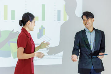 Fototapeta na wymiar Male businessman presenter holding hand showing presenting graph chart information data on wall projector screen to audience while female businesswoman colleague duo standing smiling