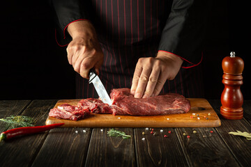 The chef cuts raw fresh beef meat on a cutting board before baking or grilling. Working environment in the kitchen of a restaurant or hotel