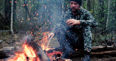 Man with cup of tea the fire in the forest. Human looks at the bonfire and drinks tea on the nature in the summer. Alone traveler sits and rests by the fire with a cup of coffee at dusk.