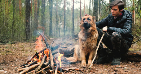 Young man with big dog sits at the fire in the forest. Cheerful guy is stroking his dog, outdoor....