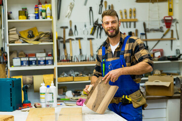 Handsome young carpenter sanding peace of wood and smiling while posing for camera