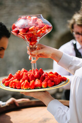 A waiter in a white shirt holds a dish of ripe strawberries with one hand and a glass vase filled with strawberries with the other hand.