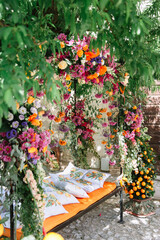 Wrought iron garden arch in an Italian garden with a bench, an orange cushion and pillows depicting tangerines. The arch is entwined with bright multi-colored flowers, in the background is a tangerine