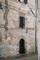 An old abandoned facade with peeling paint of an Italian house on a European street. Black wooden shutters, arched doors, old paving.