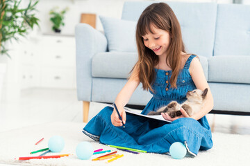 A cute girl sits on the floor at home, draws with colored pencils and plays with a little kitten. The concept of children and love for animals