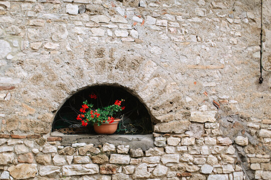In the arched niche of an old stone wall, there is a planter with red flowers. Romance of European streets.