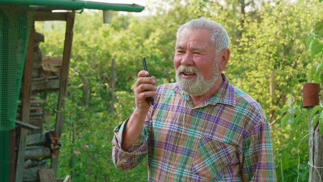 an elderly gray-haired man with a beard in a plaid shirt is talking on speakerphone on the phone. mobile communication and Internet in the village and on the farm. hearing problems in old age 