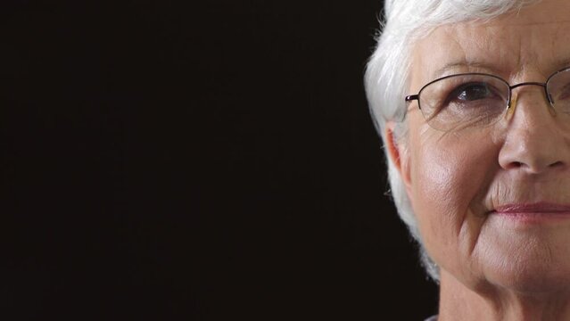 Portrait Of A Senior Woman Looking Hopeful For Humanity With Copy Space. Closeup Of A Mature Lady With Reading Glasses For Vision Correction. Face And Eyes Of A Wise Lady Against A Black Background