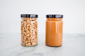 glass jars with peanuts and peanut butter on white marble background, healthy pantry ingredients