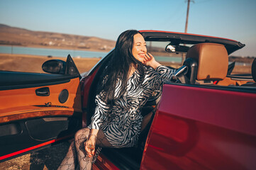 Happy smiling brunette woman driver sitting in new red cabriolet car on beach coast, drinking glass...