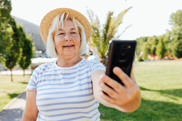 Fototapeta premium Mature blonde woman in casual summer clothes browsing smart phone while streets of city. Caucasian grandmother in straw hat holding phone and taking a selfie outdoors, summer time, technology concept