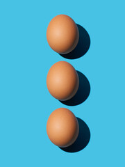 Three brown chicken eggs lie on top of each other on a blue background. Bright light, deep shadow, vertical. Close-up, top view.