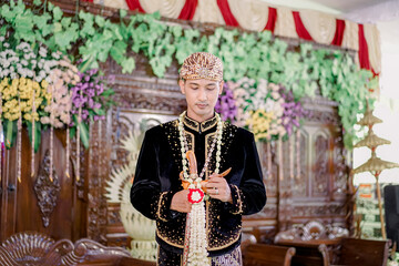 The groom wears Javanese traditional clothes, black and blangkon clothes and a jasmine necklace at...