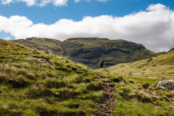 A hillwalker hiking up a hill trail with a view of a mountain. Beautiful green Scottish mountain scenery in the Trossachs National Park, summer