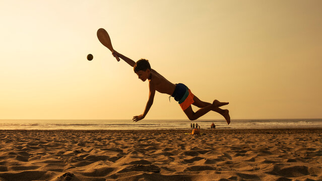 Happy Boy Playing With Ball On The Beach At Sunset. Young Man Returning The Ball By Throwing Himself On The Sand Of The Beach. Child Playing On Vacation Taking Advantage Of Last Days Without School