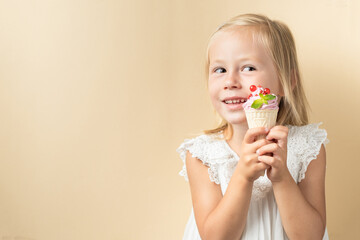 Beautiful little caucasian girl with blonde hair in a white dress enjoying berry flavored waffle cone ice cream on the beige background