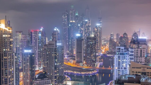 View of various skyscrapers in tallest recidential block in Dubai Marina aerial during all night timelapse with artificial canal. Many towers with blinking lights and yachts. Cloudy sky