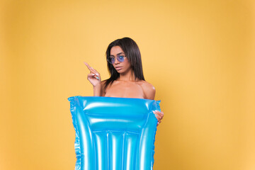 Young African American woman wearing bikini, holding an air mattress points to the side on yellow...