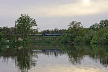 lake water with green trees on the shore and a high embankment with electric train cars on the railway against the background of a gray sky