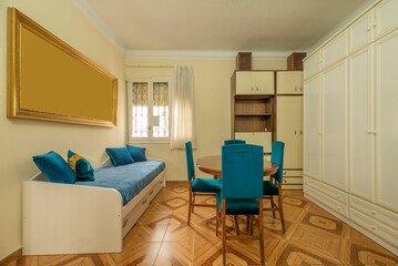 Living room of a house with a wooden dining table with blue upholstered chairs and a sofa bed with matching upholstery and kitsch vintage tile flooring and a large gold frame on one wall