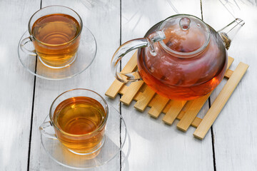 Tea in glass teapot and two cups on a wooden white background with hard shadows. Teapot with green or black tea with reflection in glass in the country. Tea time