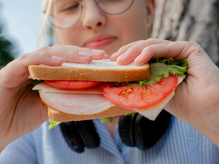 A young girl student in the park holds a juicy and tasty sandwich with sausage and tomatoes in her...
