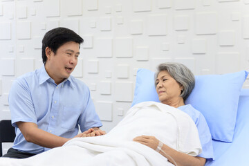 Senior Asian female patient resting on the medical bed in hospital and talking to her son. 