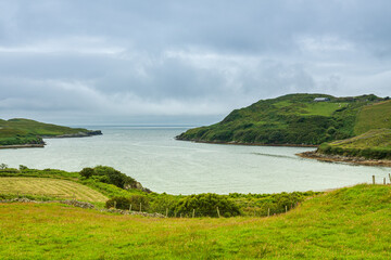 Fototapeta na wymiar A wide bay with hills covered with green trees under a thick cloudy sky. The sandy beach of a cove-shaped bay with lush green grass near the edge of the ocean.