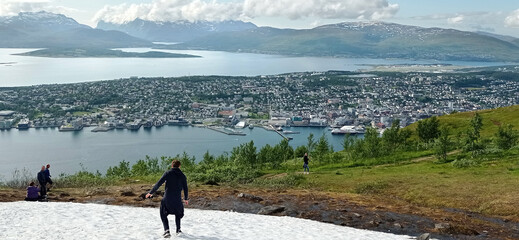Snow on the top of the Mount Storsteinen in summer and panoramic view of Tromso city in Norway. Man slides down from snowy mountain in Tromso, Norway.