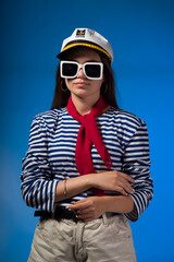 A young, attractive girl with long, straight hair, brown eyes in a vest, a captain's cap and a red neckerchief on a blue background looks into the distance through dark glasses. Close-up portrait. 