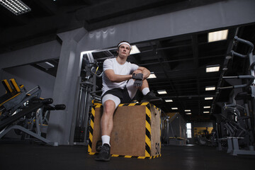 Fototapeta na wymiar Low angle full length shot of a handsome athletic man sitting on a jumping box at gym, resting after exercising