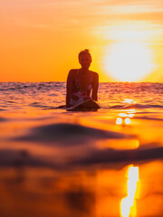 Silhouette of surfer woman on a surfboard in ocean with sunset or sunrise tones.