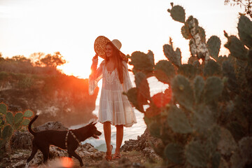Girl in a hat and a white dress walks a dog on the shore with cacti overlooking the ocean and sunset.