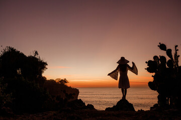 Woman in a dress and a straw hat stands on top of a rock near a cactus and looks at the sea at sunset
