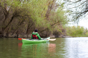 Young woman in green kayak paddle at river near trees with gentle green leaves at spring