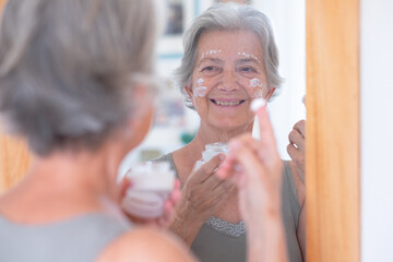 Close up shot of senior beautiful woman applies anti aging cream on wrinkled face - elderly smiling lady holding a cosmetic jar product
