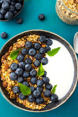 Homemade granola with yogurt, blueberries and mint leaves in a bowl on a turquoise background, delicious healthy breakfast
