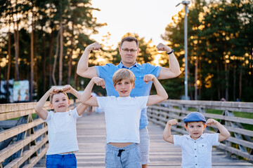 Portrait og happy family - father and his three children sons smiling and showing their muscles outdoor in park on background. Two different generation concept.