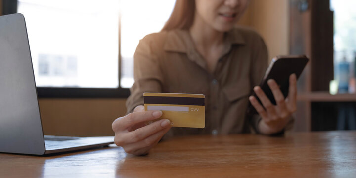 Closeup image of a woman's hand holding credit card and pressing at mobile phone on wooden table in office