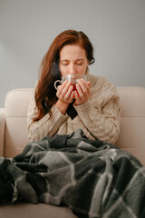 Woman covered herself with a blanket while sitting on the couch drinking hot tea for illness.
