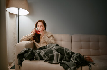 Young woman caught a cold by wrapping herself in a blanket at home and drinking hot tea from a cup.